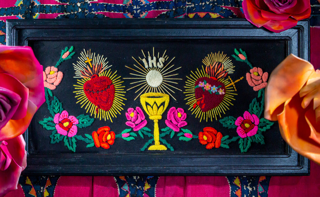 Embroidery of two brightly colored bleeding hearts flanking a monstrance amid a garland of large flowers in reds and pinks.